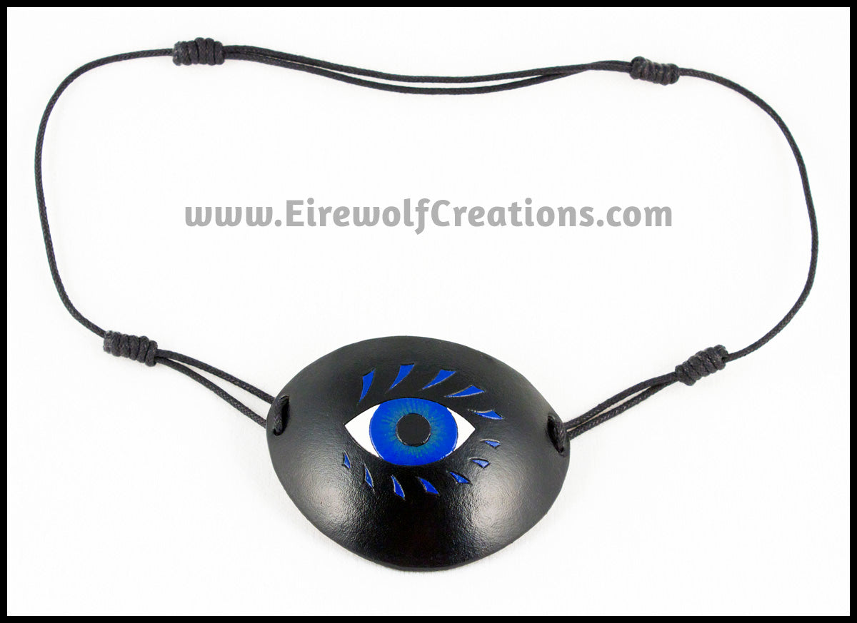 A bold graphic stylized blue eye with blue eyelashes carved and painted onto a handmade black leather eye patch, for a masquerade costume or pirate cosplay. By Erin Metcalf of Eirewolf Creations.