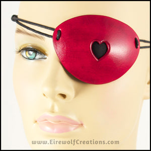 A handmade leather dyed red eye patch with a heart cut out, backed with transparent black fabric to allow some visibility, for a masquerade costume or pirate cosplay. By Erin Metcalf of Eirewolf Creations.