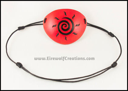 A handmade leather eye patch dyed orange with a bold black sun spiral symbol carved and painted on the leather, for a masquerade costume or pirate cosplay. By Erin Metcalf of Eirewolf Creations.