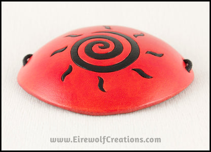 A handmade leather eye patch dyed orange with a bold black sun spiral symbol carved and painted on the leather, for a masquerade costume or pirate cosplay. By Erin Metcalf of Eirewolf Creations.